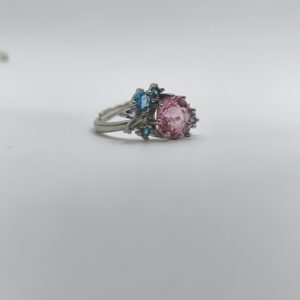 نمای نزدیک از انگشتر طرح گل با نگین صورتی و نگینهای آبی و بنفش روی زمینه سفید.