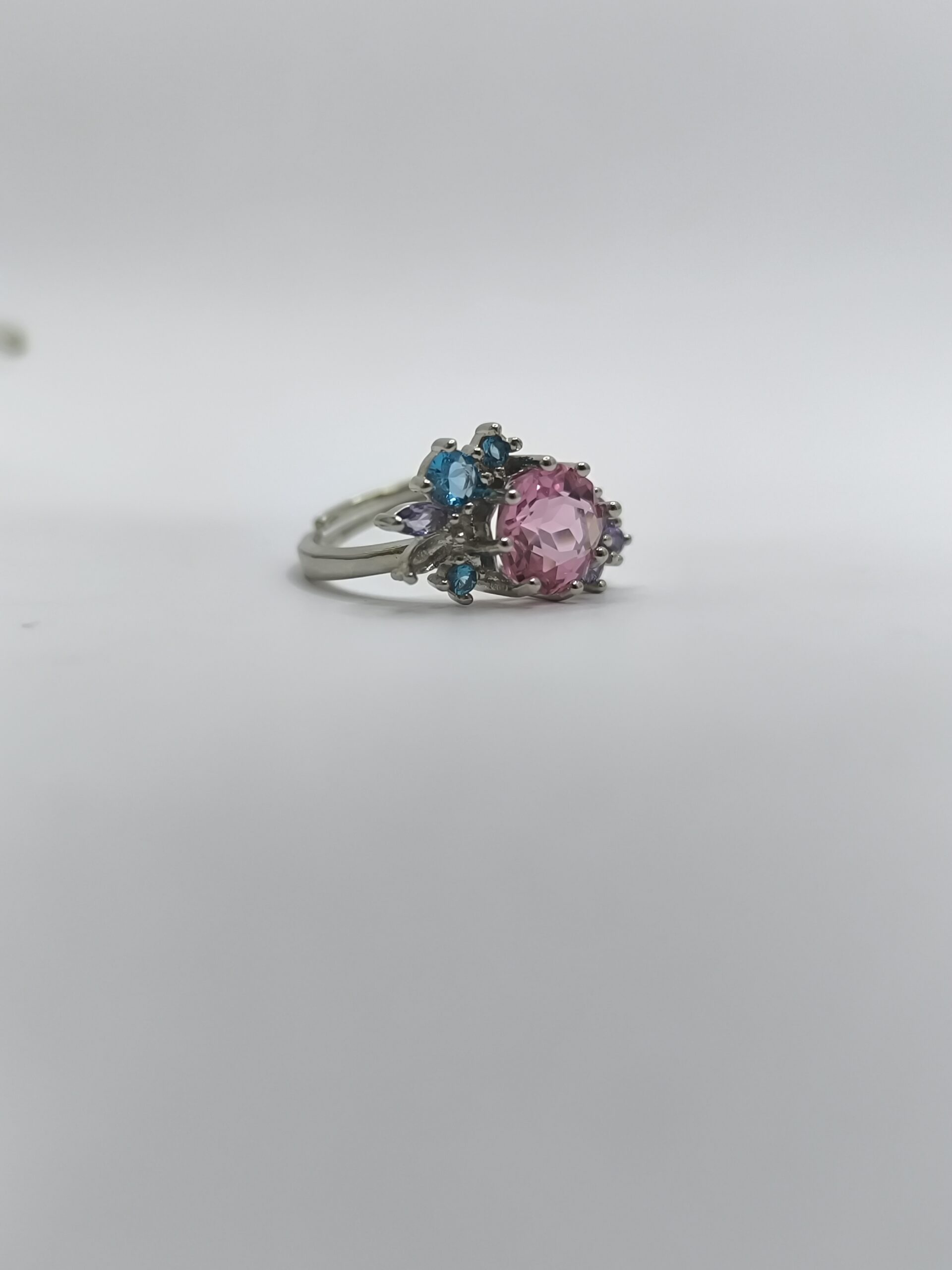 نمای نزدیک از انگشتر طرح گل با نگین صورتی و نگینهای آبی و بنفش روی زمینه سفید.