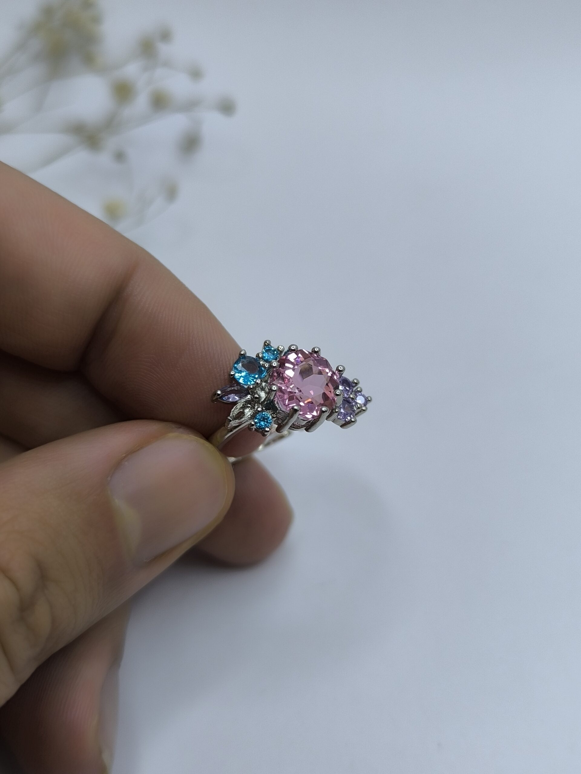 نمای نزدیک از انگشتر طرح گل با نگین صورتی و نگینهای آبی و بنفش روی زمینه سفید.