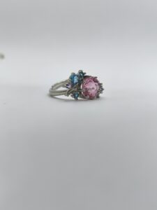 نمای نزدیک از انگشتر طرح گل با نگین صورتی و نگین‌های آبی و بنفش روی زمینه سفید.